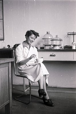 A seated woman in a lab coat examines a petri dish, appearing thoughtful. Chrome chair and lab equipment (beakers, balances) ...