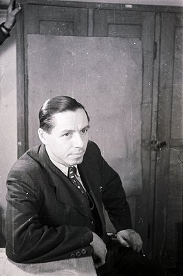 Formal portrait of a man in a suit and tie, seated with hands clasped. Sparse background features a closed wooden door. Likel...