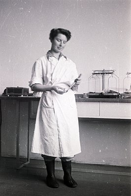 A young woman in a lab coat and tall black boots examines a small vial. Likely a chemist or researcher, she stands in a labor...