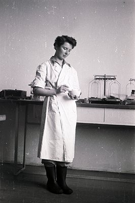 A woman in a lab coat and dark boots stands near a lab bench examining an object in her hands. Apparent period details (equip...