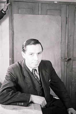 A seated man in a suit and tie looks directly at the camera. He appears thoughtful, positioned behind a table against a plain...