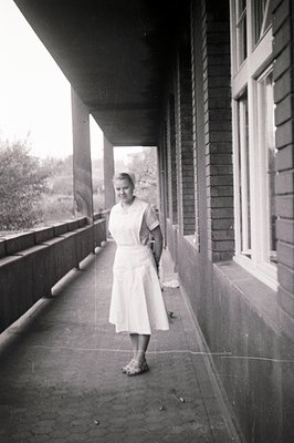 A young woman in a nursing uniform stands on a covered porch with wooden details. The exterior view reveals greenery and a bl...