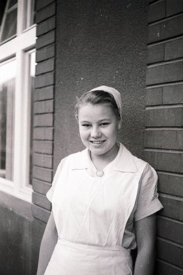 Young nurse in uniform and head covering, posed for a portrait. Classic mid-century style, likely 1950s. Textured brick wall ...