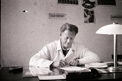 Man in a white coat signs a document at a desk, illuminated by a vintage desk lamp. A sign reading "Zdarovye" is visible on t...