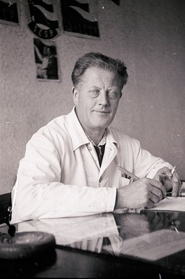 Man in a lab coat, seated at a desk with papers, pen in hand. Appears to be a scientist or researcher. Posters and signs visi...