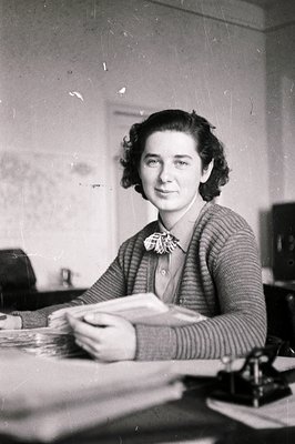 A seated woman, likely an office worker, smiles directly at the camera. She wears a cardigan over a collared blouse with a sm...