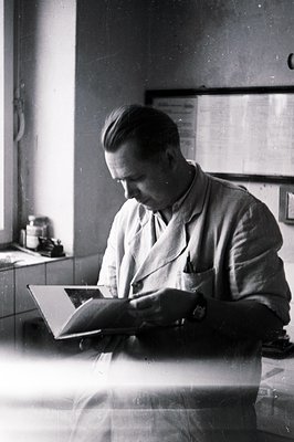 A man in a lab coat studies a book, seated at a tiled counter in a clinical setting. Visible details include a clock, glasswa...