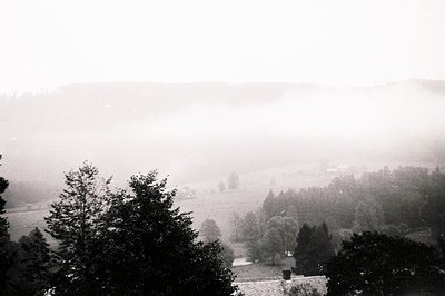 Ethereal landscape view, thick fog obscures valley floor. Partially visible rooftops suggest a village or small town nestled ...