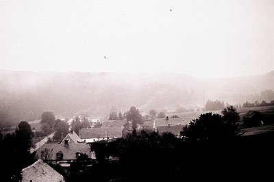 A monochrome landscape view showing a village nestled within a valley. Buildings with sloped roofs are visible, surrounded by...