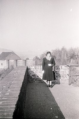 A woman in a dark coat and hat stands on a stone retaining wall overlooking a valley with a village in the distance. Likely 1...