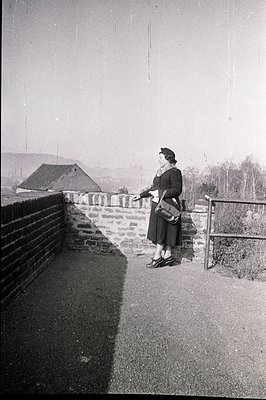 A woman in a tailored coat and hat poses near a brick wall and metal railing, overlooking a view with rooftops and distant tr...