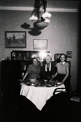 A formal interior scene shows three people seated around a table laden with food. The wallpapered room includes framed artwor...