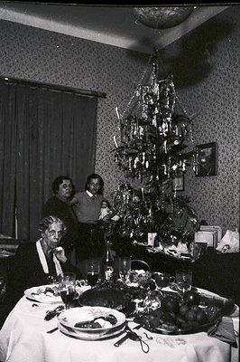 Festive gathering around a table laden with food and a tall, ornate Christmas tree. Three figures are present, two women and ...