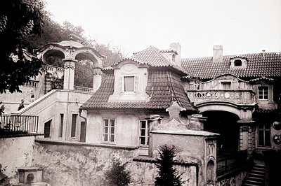 A grand, multi-level villa features a central gazebo, ornate balconies, and weathered stone detailing. A small figure stands ...