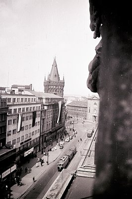 Elevated view of a bustling city street, likely Prague, featuring a distinctive spire and trams. Buildings display signage, h...