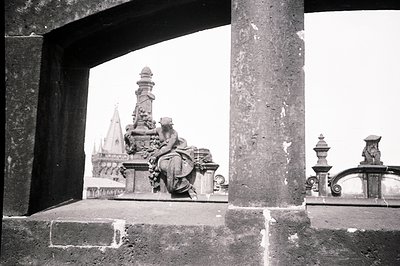 View framed by architectural details reveals a sculpted rooftop scene. A bronze figure, likely a historical personage, reclin...