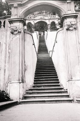 Ornate, monumental stone staircase ascends toward a balustraded landing & arched gateway. Formal garden visible beyond. Possi...