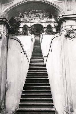 Monumental, weathered stone staircase ascends toward a lushly planted balcony and arched facade. Ornate carvings embellish th...