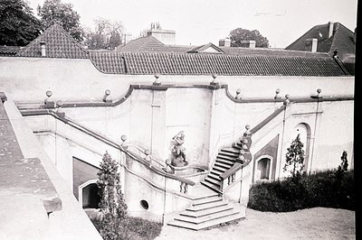 A formal garden features a grand, terraced staircase leading to a building with red-tiled roofing. Sculptural details and arc...