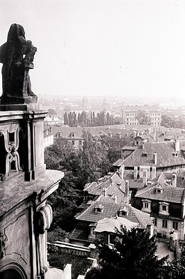 High-angle view of rooftops in an older European city, likely Prague, Czech Republic. A stone statue and decorative architect...