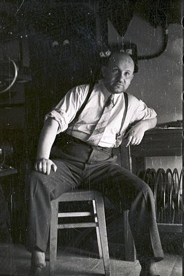 A seated man, likely a craftsman or artist, wears a collared shirt and suspenders; he leans against a work table with visible...