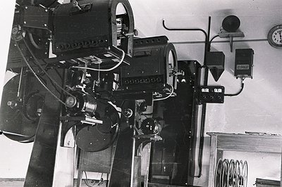 A large, vintage 35mm film projection system dominates the frame. Likely from the 1950s or 60s, the machine features a comple...