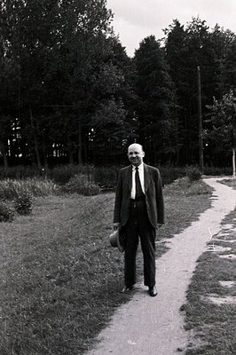 Formal portrait of a balding man in a suit and tie, holding a hat, standing on a winding pathway through a wooded area. Likel...