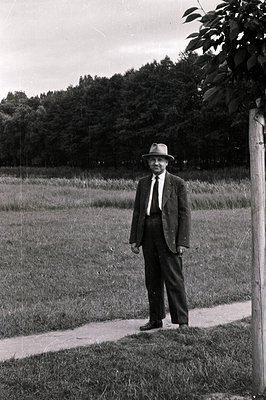 Formal portrait of a man in a suit and hat standing near a pathway and trees. The subject's attire suggests a professional or...