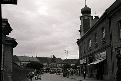 A view of a European town street scene with a prominent church façade and shopfront signage. Several pedestrians and vintage ...