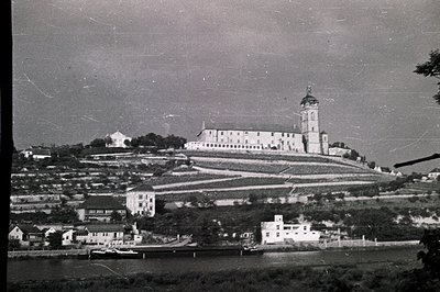 A grand, tiered building with a central spire dominates this hillside landscape. Terraced vineyards slope toward a riverbank ...