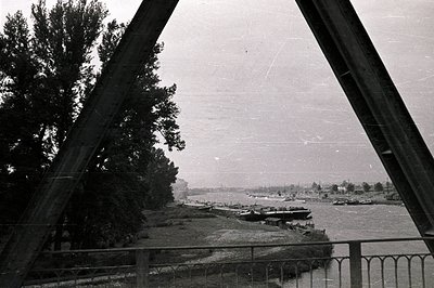 Framed view from a bridge overlooking a broad river; numerous barges are moored along the riverbank. Lush greenery lines the ...
