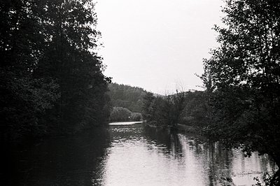 A serene, tranquil river scene framed by dense foliage. The waterway reflects the overcast sky and surrounding trees, creatin...