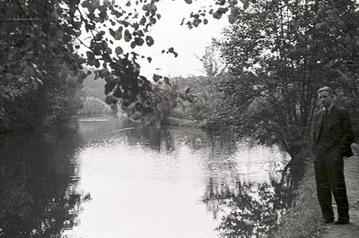 A formally-dressed man stands beside a reflective body of water, likely a lake or wide river, framed by dense trees. The imag...