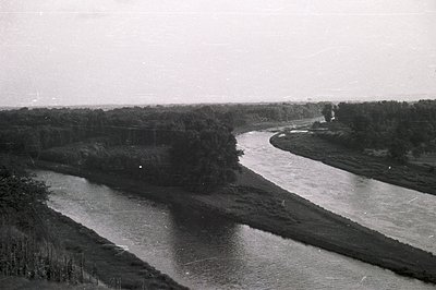 A wide, gently curving river winds through a flat, grassy landscape. Dense riparian vegetation lines the banks. A small, simp...