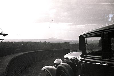 Classic 1930s automobile parked beside a curved brick retaining wall. Distant, rolling hills and a solitary peak dominate the...