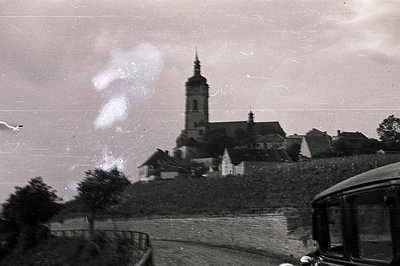 A high-angle, grainy view captures a church steeple rising above a village landscape. Stone buildings and rooftops are visibl...