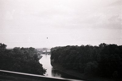 Monochrome view of a river winding through dense trees. A bridge spans the waterway, receding into distance. Likely a roadsid...