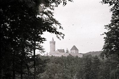 A dramatic, high-angle view frames a medieval castle set within a dense forest landscape. The castle features a prominent tow...