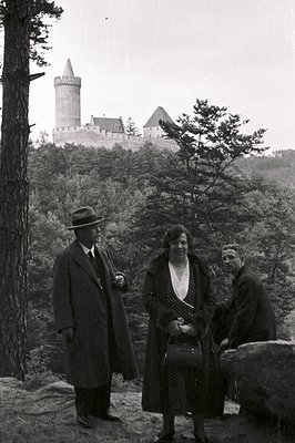 Three figures pose before a large stone castle perched atop a forested hill. The individuals are formally attired: a man in a...