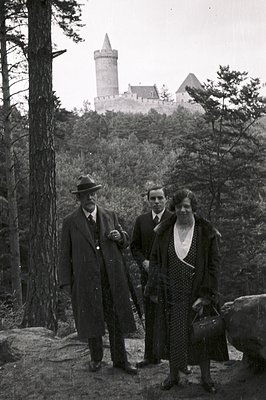 A formal portrait showcases a man in a fedora, a younger man, and a woman with a vintage handbag, posed before a medieval cas...