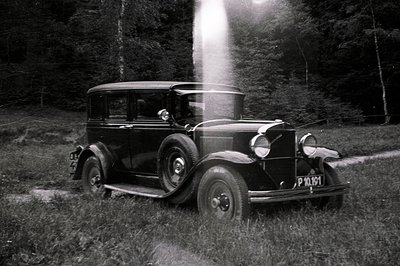 A black 1930s sedan sits on a grassy verge beside a dirt road, backed by a dense forest. The vehicle's license plate is legib...