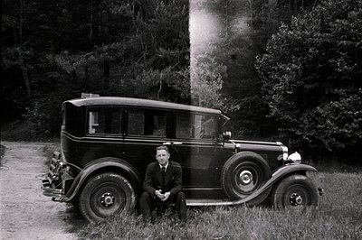 A man in a dark suit sits beside a classic 1930s sedan, likely a Durant. The car’s simple design and tire details are charact...