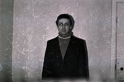 Man in a dark suit and turtleneck stands close to textured wall with a door visible right. Minimalist portrait, stark lightin...