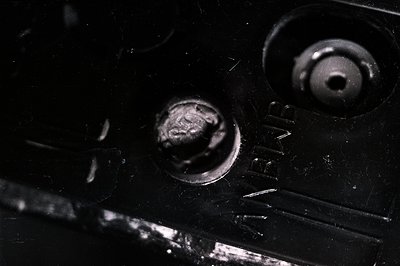 Close-up of weathered, textured metal surface. Two corroded screw heads are visible, one significantly rusted, set within a d...