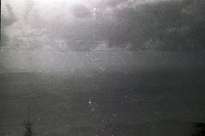 Dramatic, overcast sky dominates this monochrome photograph. Visible film grain and numerous scratches suggest age and potent...