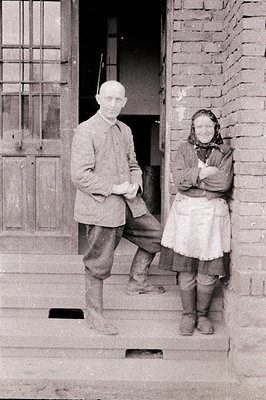 A man and woman pose outside a brick building, likely a farm or rural residence. The man wears a suit jacket, knickerbockers,...