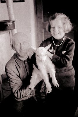 A man and young girl pose indoors, the girl holding a baby goat aloft. The man wears a long-sleeved shirt; the girl, a dark s...