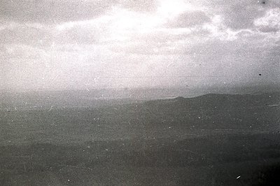 A monochrome landscape showing a distant shoreline and cloudy sky. Visible grain and surface scratches suggest an older photo...