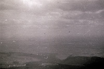 Monochromatic landscape showing a hazy horizon with low, distant hills and a cloudy sky. Extensive scratches & dust artifacts...