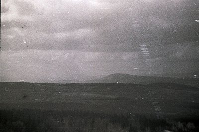 Striking monochrome landscape. Rolling hills lead to a distant mountain range under a heavily clouded sky. Visible film grain...